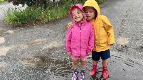 two young children in hooded rain jackets stand in a rain puddle together