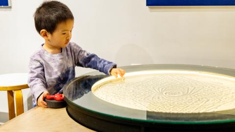 a small boy looks at a glass-topped table filled with a sand design