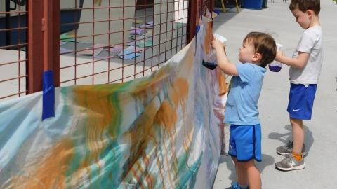 A white sheet full of paint splatter is taped to a fence where two boys are spraying it with paint from spray bottles.