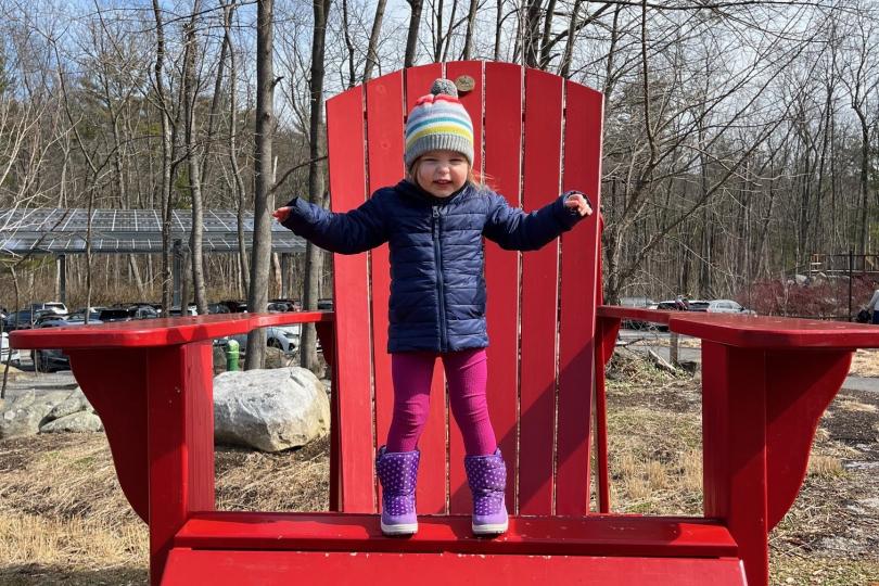 a small girl stand on the seat of a giant red Adirondack chair